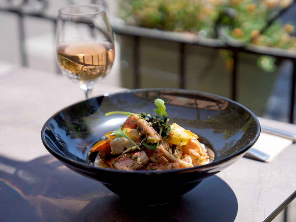 Salade et viande en tranches servies en terrasse, accompagnées d’un verre de vin blanc, dans un décor végétal.