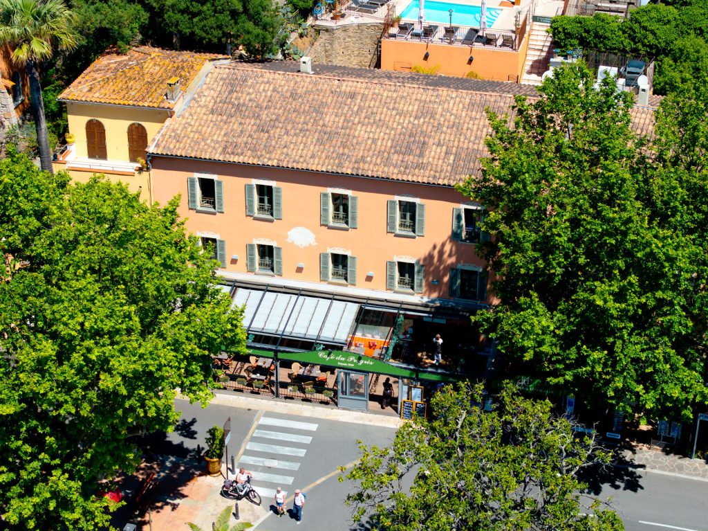 Vue aérienne du restaurant Le Progrès, avec sa façade couleur abricot, ses volets verts et sa terrasse sous les arbres.