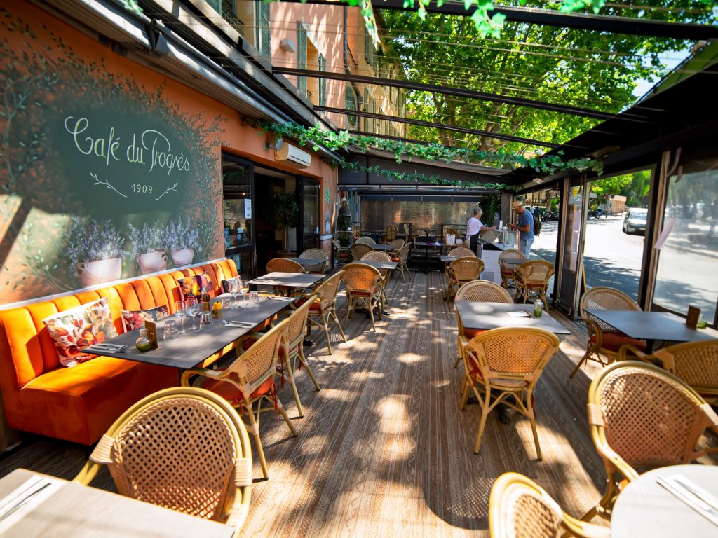 Terrasse historique du Progrès, fondée en 1909, avec ses chaises en osier et son banc orange, au milieu d'une végétation luxuriante.