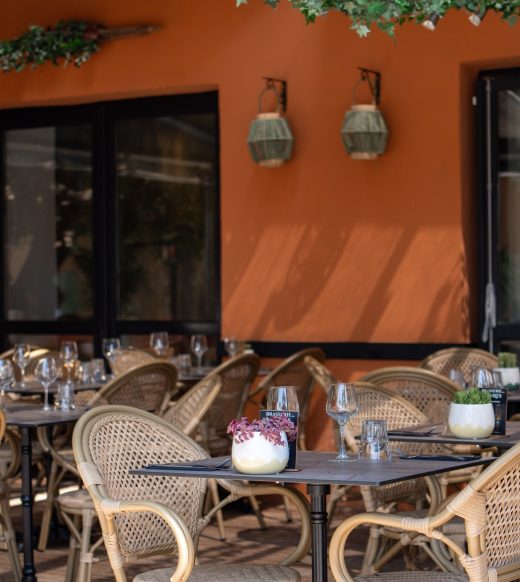 Terrasse historique du Restaurant Le Progrès, chaises en osier, vaisselle, fleurs et lanternes sous verdure.