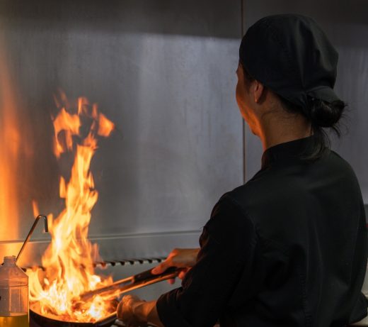 Chef en noir flambe un plat au gaz, perpétuant la tradition culinaire du Restaurant Le Progrès.