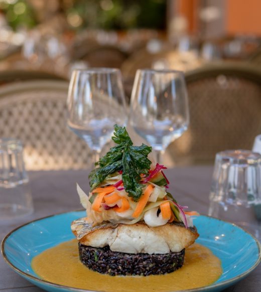 Poisson sur riz noir et légumes, dressé à table, deux verres vides, terrasse en fond.
